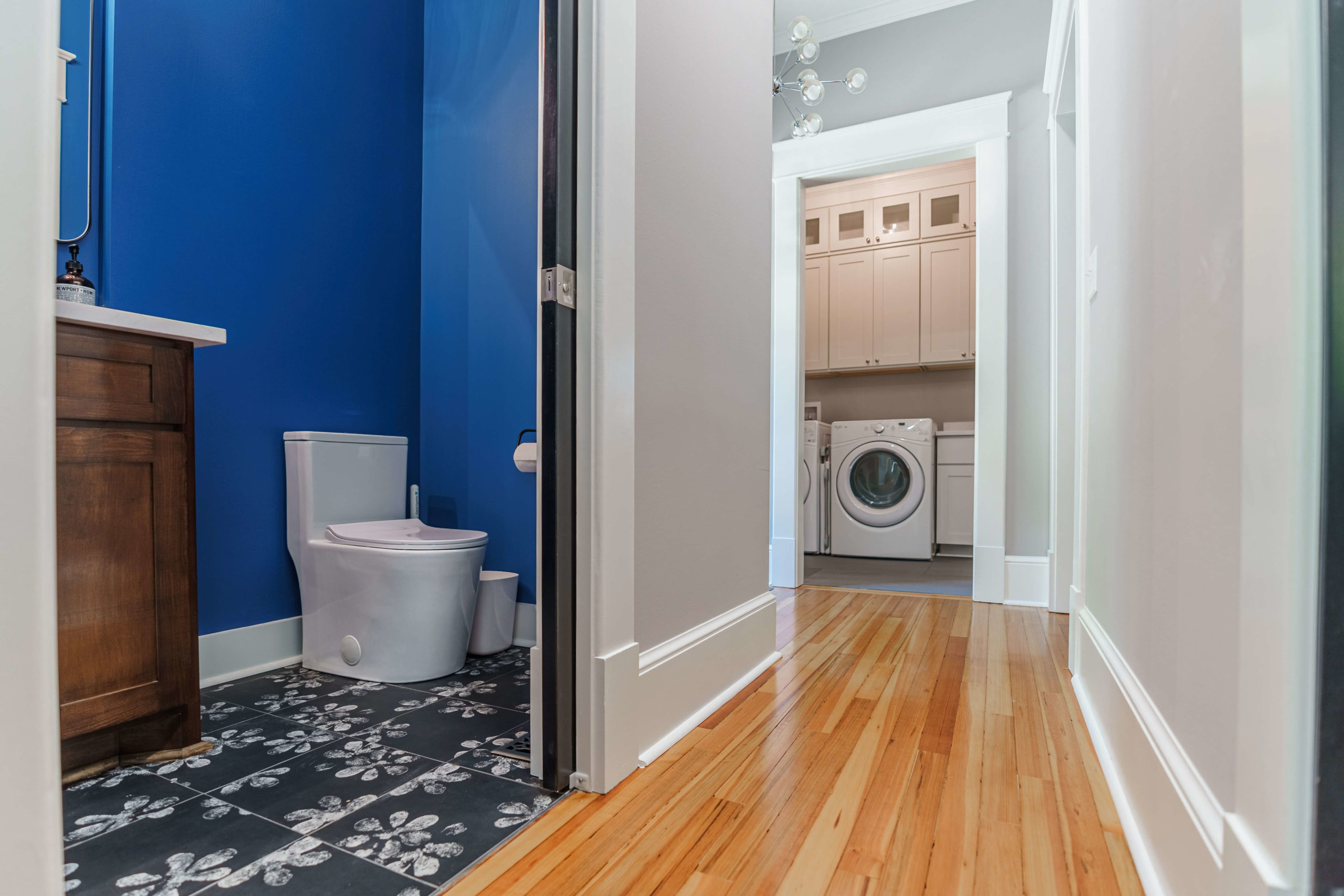 Wood floors in a hallway that leads the the laundry room and the half bath with blue walls and floral pattern tile